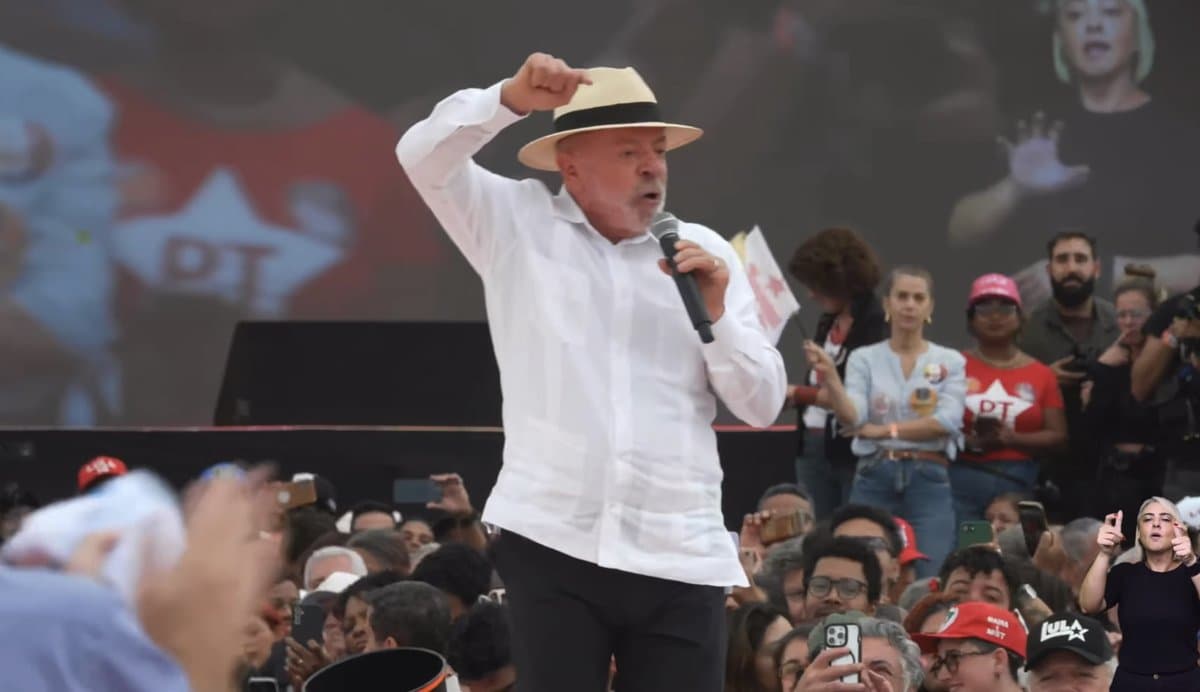Presidente Lula discursa com microfone na mao durante comicio, cercado por apoiadores com bandeiras do PT