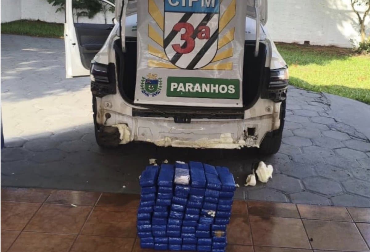 Tabletes de maconha empilhados no chão atrás de uma viatura da 3ª CIPM da Polícia Militar em Paranhos, no Mato Grosso do Sul, com o para-choque removido