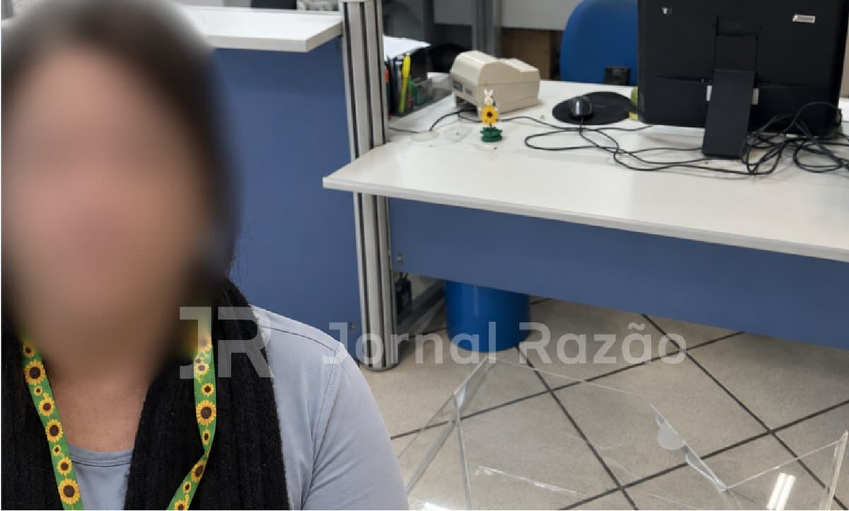 Mulher com rosto desfocado diante de guichê do INSS com acrílico quebrado no chão, em Joinville