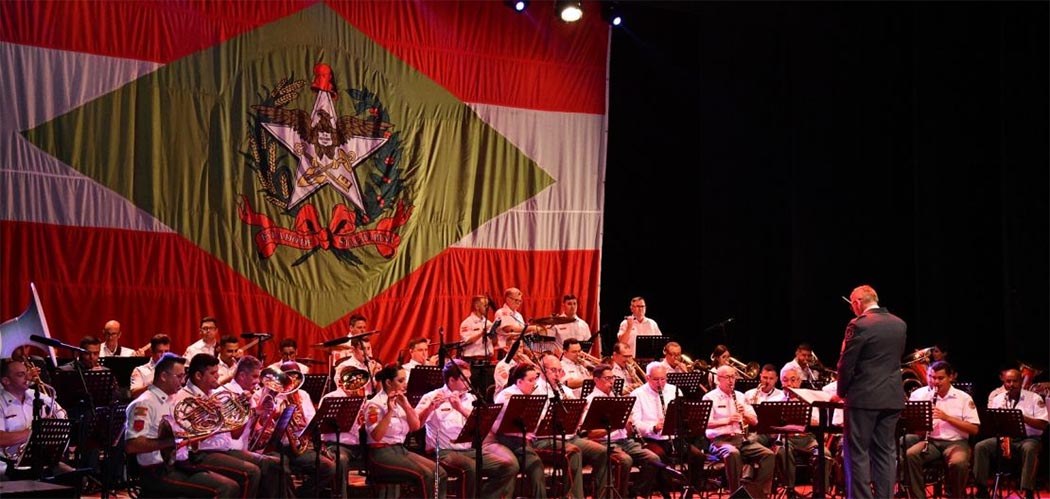Banda de Música do Corpo de Bombeiros Militar de Santa Catarina se apresenta em palco com bandeira de SC ao fundo