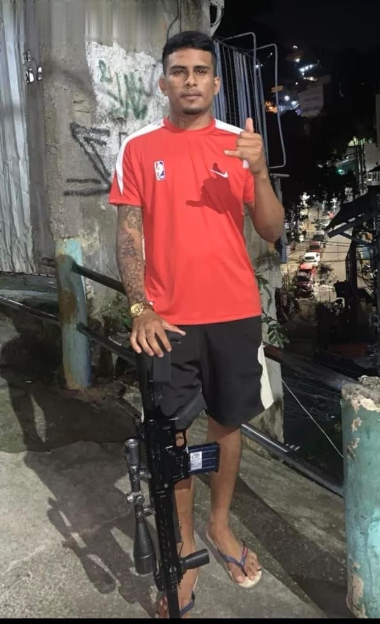 Homem de camiseta vermelha posa ao lado de fuzil apoiado em corrimão de uma laje, à noite, em comunidade do Rio de Janeiro