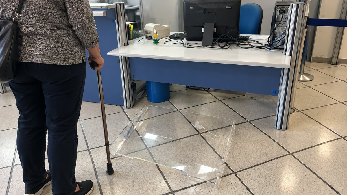 Proteção de acrílico do guichê do INSS caída no chão diante da mesa de atendimento, em Joinville